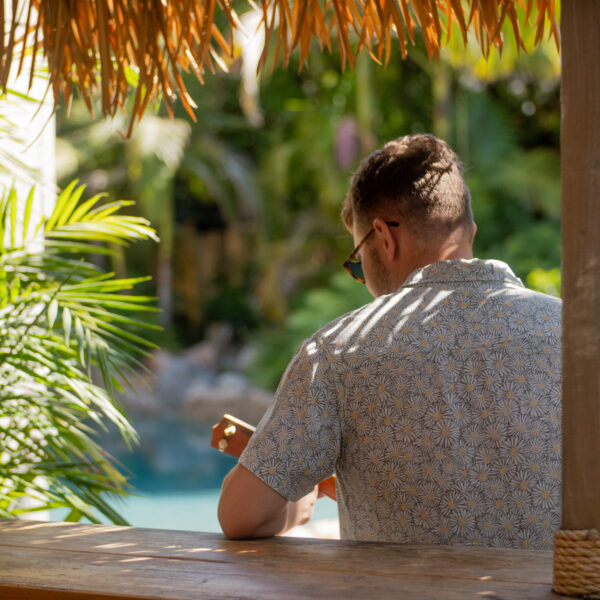 Talent playing ukulele, sitting at bar counter with Lagoon pool and palms in background at Tropic Shots production set