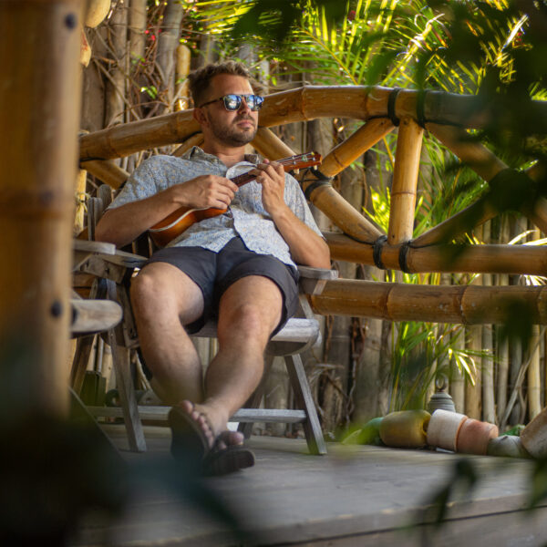 Talent sitting in Palapa playing ukulele with dense jungle foliage backdrop at Tropic Shots production set